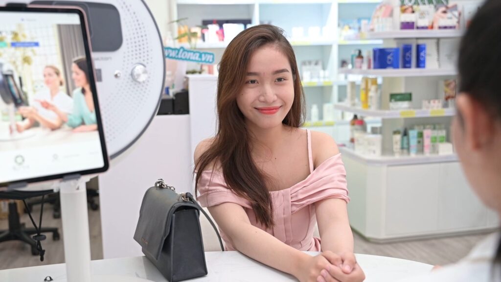 Khách ngồi bên máy soi da OBSERV tại khu tư vấn, phía sau là kệ sản phẩm — Phòng khám Chuyên khoa Da liễu Lona Skin Lab, Cần Thơ