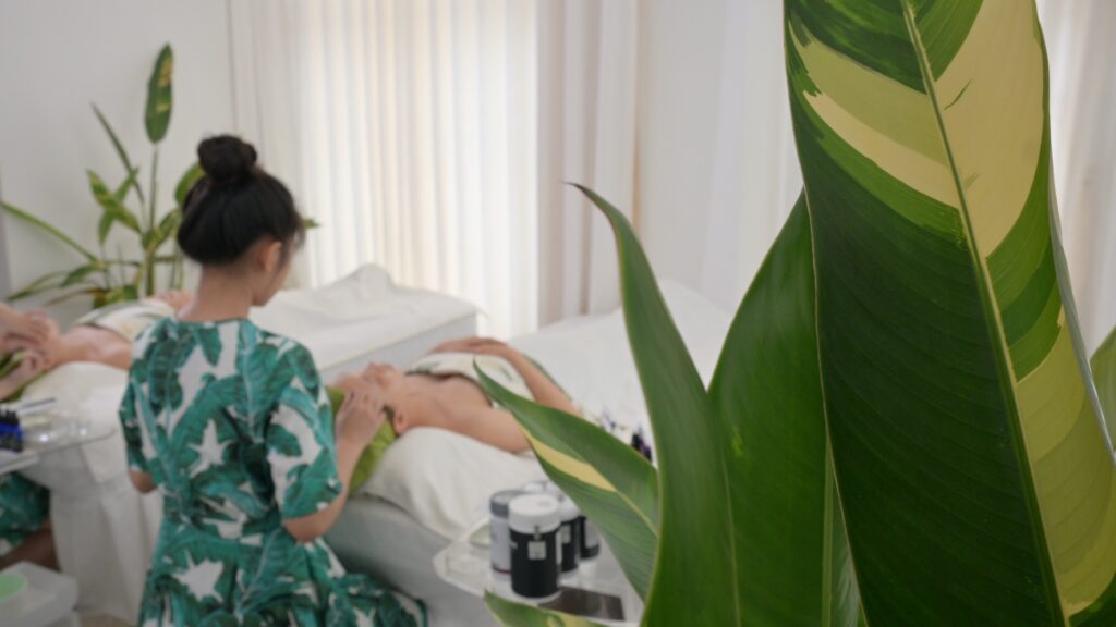Kỹ thuật viên mặc đồng phục hoạ tiết lá chuối chuẩn bị giường trị liệu, bên cạnh là cây xanh — Phòng khám Chuyên khoa Da liễu Lona Skin Lab, Cần Thơ
