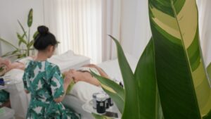 Kỹ thuật viên mặc đồng phục hoạ tiết lá chuối chuẩn bị giường trị liệu, bên cạnh là cây xanh — Phòng khám Chuyên khoa Da liễu Lona Skin Lab, Cần Thơ